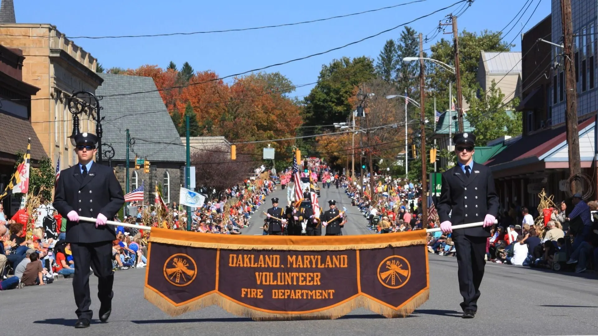 Two Oakland Volunteer Fire Department members carrying the department's banner in the Autumn Glory Grand Feature Parade, with spectators lining a street under fall trees
