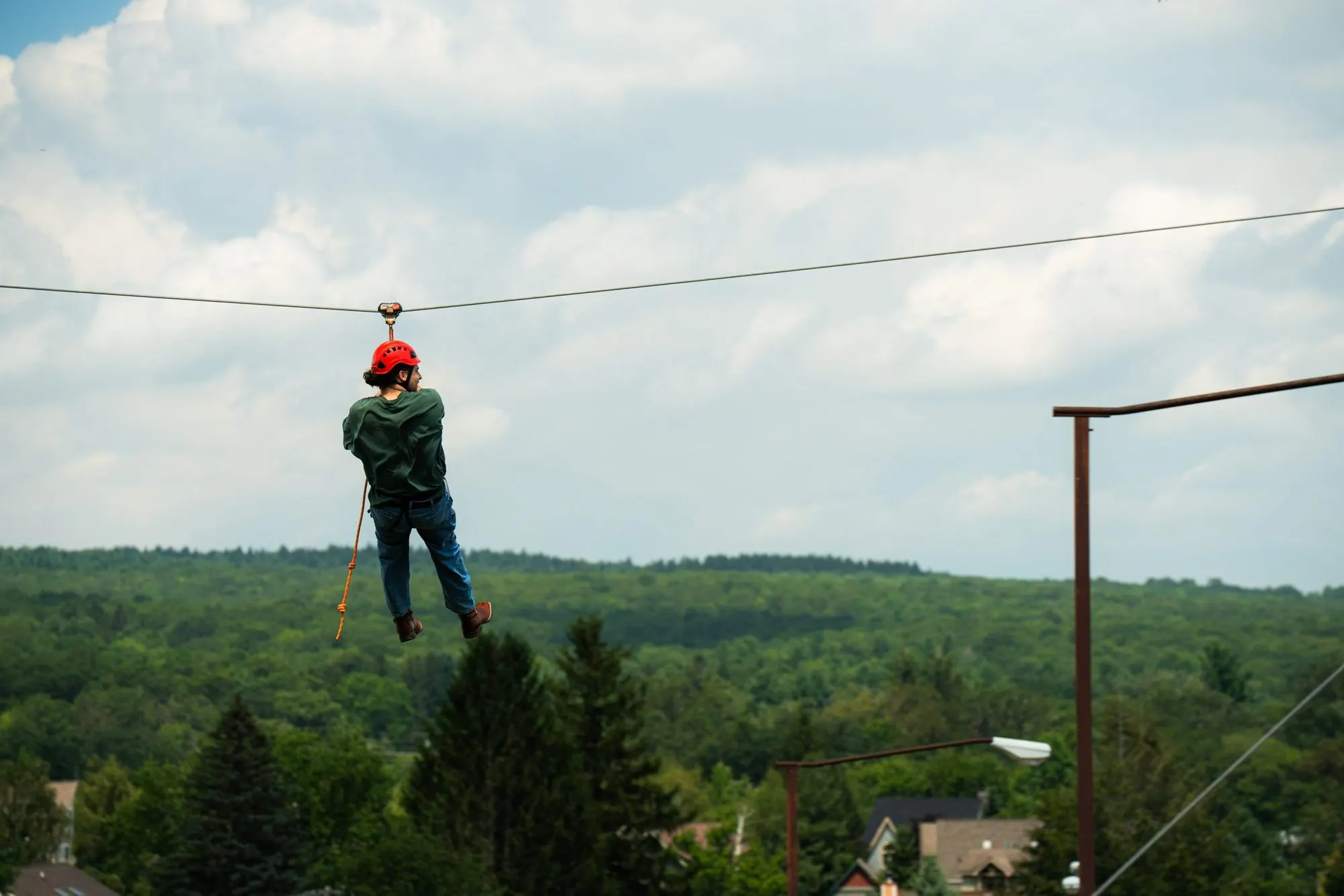 A rider in a red helmet on a zip line at Wisp Resort, above treetops and rooftops with forested ridges beyond